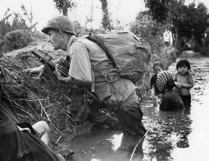 FILE - In this Jan. 1, 1966 file photo, a Paratrooper of the 173rd U.S. Airborne brigade crouches with women and children in a muddy canal as intense Viet Cong sniper fire temporarily pins down his unit during the Vietnamese War near Bao trai in Vietnam. Filmmaker Ken Burns said he hopes his 10-part documentary about the War, which begins Sept. 17, 2017 on PBS, could serve as sort of a vaccine against some problems that took root during the conflict, such as a lack of civil discourse in America. (AP Photo/Horst Faas, File)