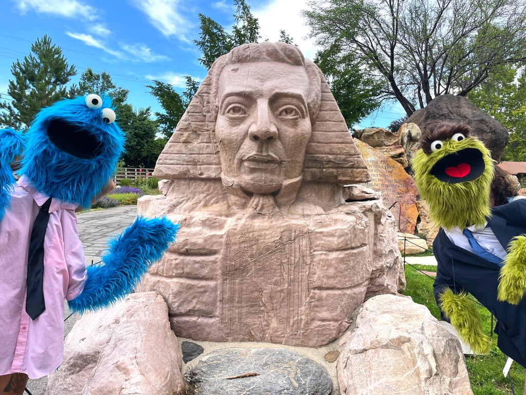 (Salt Lake Acting Company) Two of the puppet characters from "SLAC's Summer Show: A Beautiful Day in the Neighborhood" — a for-adults play looking at today's headlines through the lens of educational television — visit the Joseph Smith sphinx at Salt Lake City's Gilgal Gardens. The play runs July 12 to August 20 at Salt Lake Acting Company.
