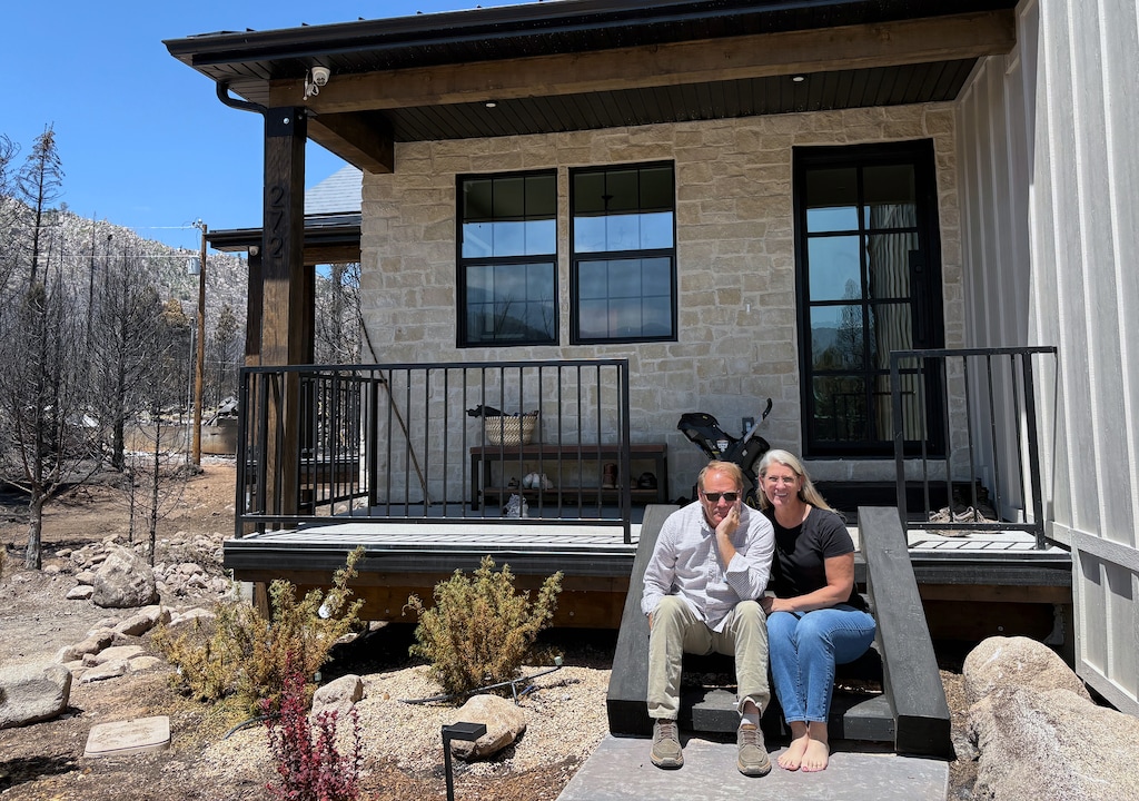 A man and woman sit on a porch of a newly-constructed home. In the background are burnt trees from the Forsyth Fire.