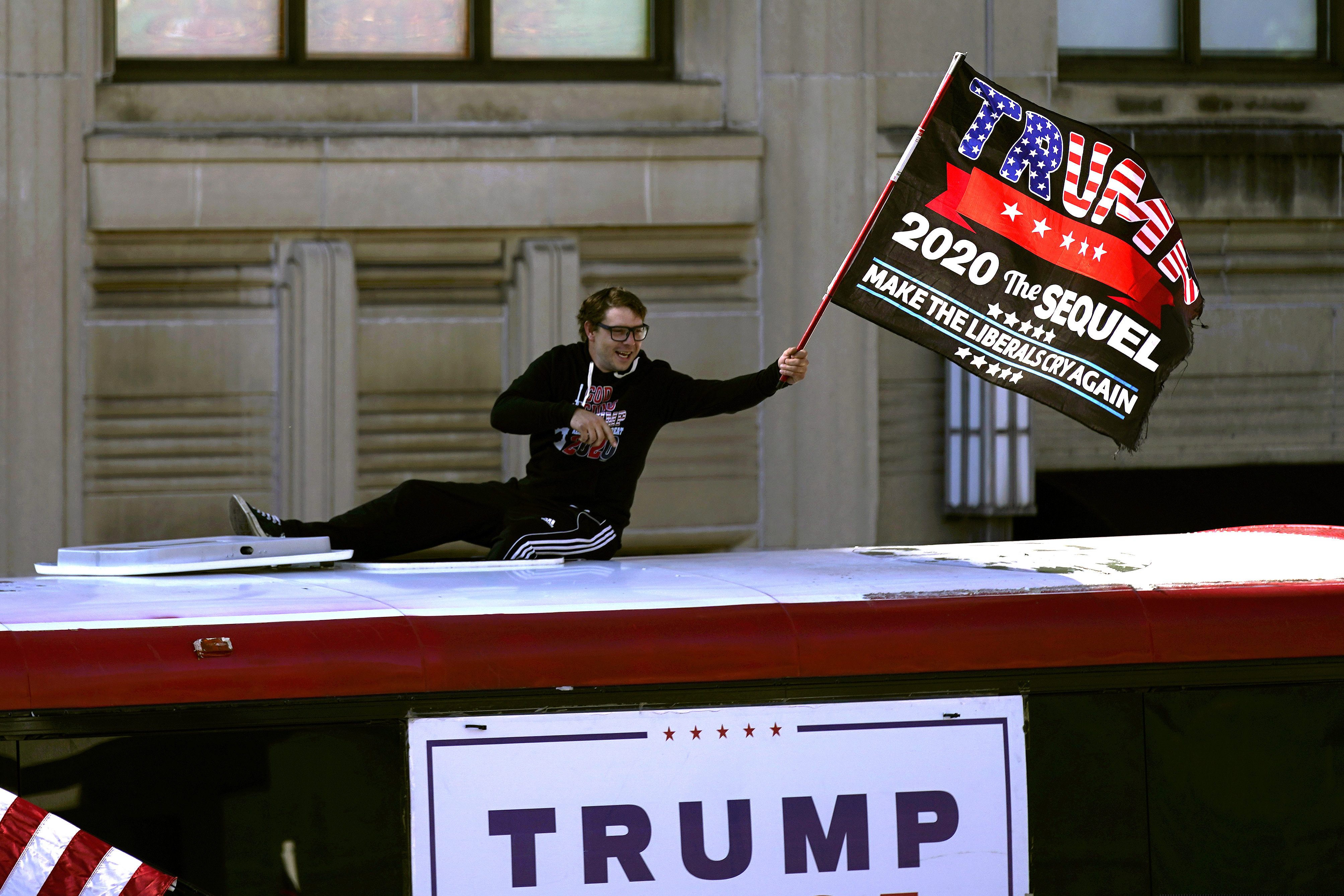 A supporter of President Donald Trump waves a flag from atop a bus outside the Pennsylvania State Capitol, Saturday, Nov. 7, 2020, in Harrisburg, Pa., after Democrat Joe Biden defeated President Donald Trump to become 46th president of the United States. (AP Photo/Julio Cortez)