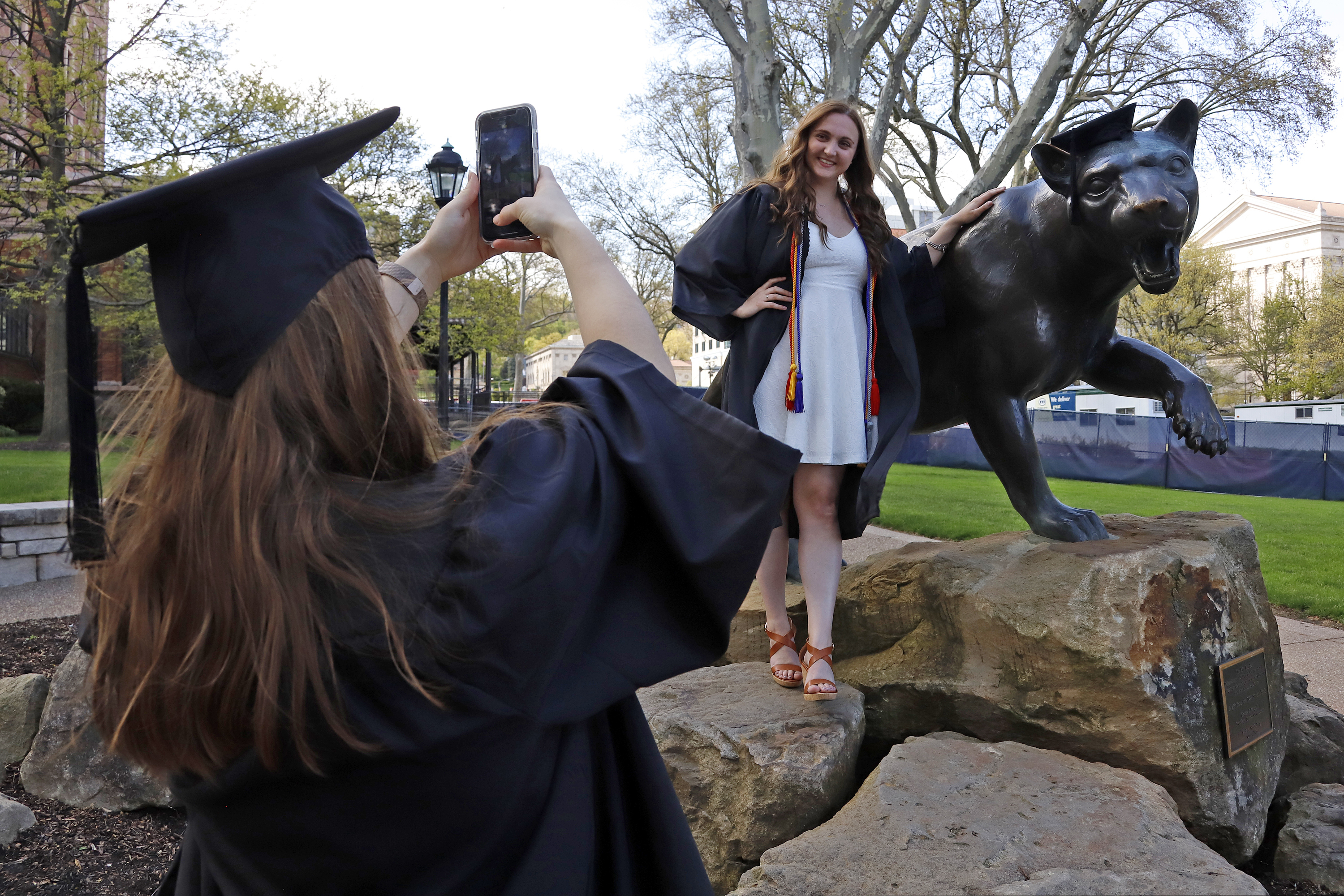 FILE - In this April 27, 2020, file photo, Class of 2020 University of Pittsburgh graduates Shannon Trombley, left, of Philadelphia, and Julie Jones, of West Chester, Pa., take turns posing for photos with a statue of Pitt's mascot, the Pitt Panther. As the coronavirus leaves colleges holding “virtual graduation” events online, many schools are recruiting famous figures to deliver speeches over their laptops instead of the lectern.  (AP Photo/Gene J. Puskar, File)